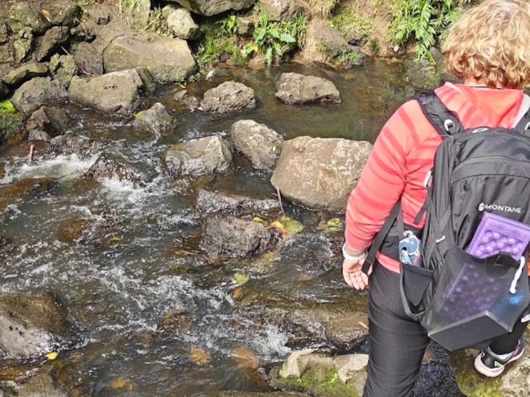 A walker with a daypack crossing a rocky stream at Oakley Creek Waterfall in Auckland, a handy local walk for building up to a walking holiday.
